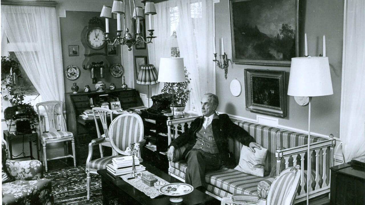 Man sitting on a Gustavian style sofa. Landscape painting above the sofa. Flowers, lamps and other ornaments are visible all around.