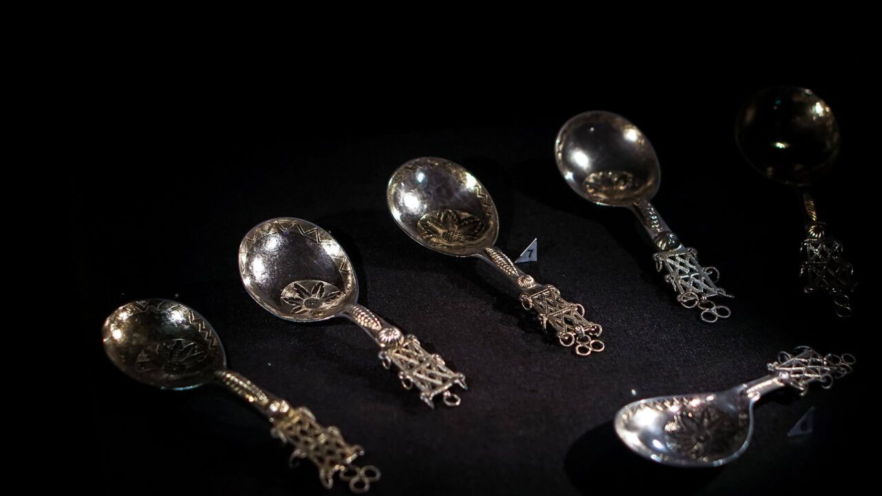 From the Silvermuseet's silver exhibition: Six silver spoons against a black background. The spoons are engraved, the handles have an openwork pattern and have rings attached.