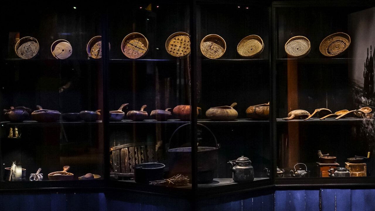 Parti ur Silvermuseets utställning Mujttalus. Stor glasmonter med nio runda träformar på hyllan högst upp. I mitten står ett flertal náhppe, mjölkstävor, samt några slevar. Längst ner bland annat grytor, kaffepannor och kaffekvarnar.