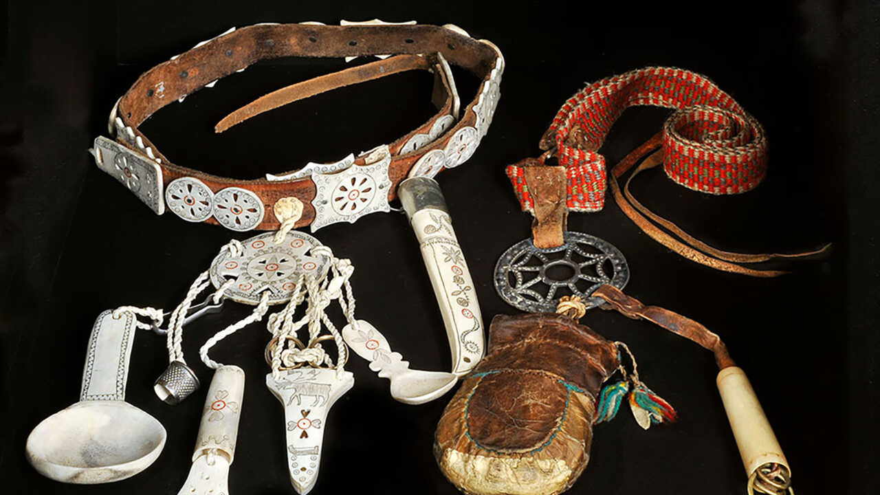 Ur Silvermuseets utställning Mujttalus: Bälte av läder med graverade hornknappar. I bältet hänger ett gehäng med kosa, fingerborg, nålhus, sax, sked och kniv. Både gehängplattan och föremålen har graverats. Bredvid ligger ett vävt bälte med röd botten med ett gehäng av metall. Gehängplattan påminner om ett spindelnät och har en skinnpåse och ett nålhus fäst vid sig.