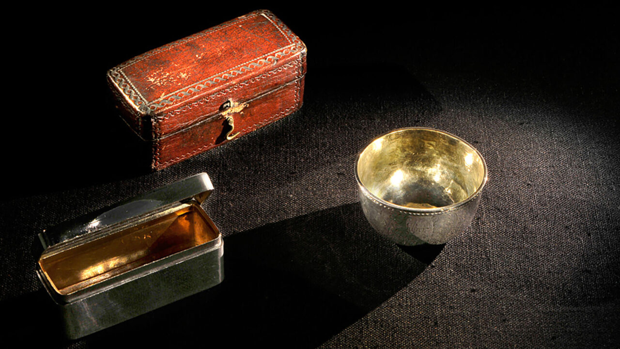 A silver rectangular small box with an open lid. Behind it is a reddish case of the same size, the case is patterned. To the right is a silver small cup.