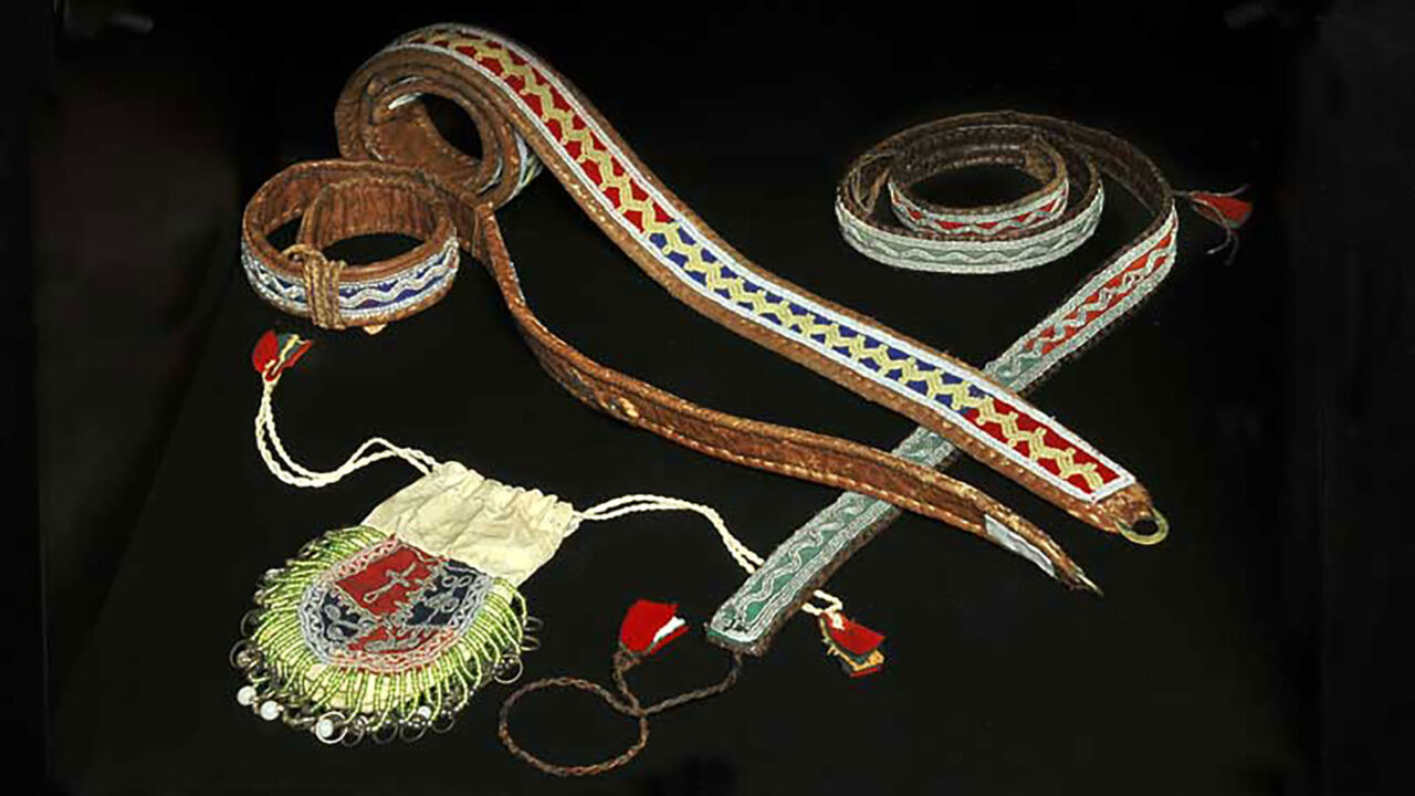 Ur Silvermuseets utställning Mujttalus: Tre bälten tillhörande samiska dräkter. Bältena har partier i rött och blått eller rött och grönt, baksida och kantning av skinn. På bältena är det sytt sicksack-mönster i tenn eller pärlor. Med på bilden syns en påse av vitt skinn, kläde, pärlor och tenn.