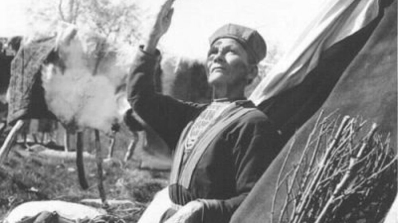 Svartvit bild där Inga Steggo står i öppningen till en tältkåta med högra armen höjd mot himlen. På sig har hon kolt och koltmössa i bakgrunden hänger flera renskinn.