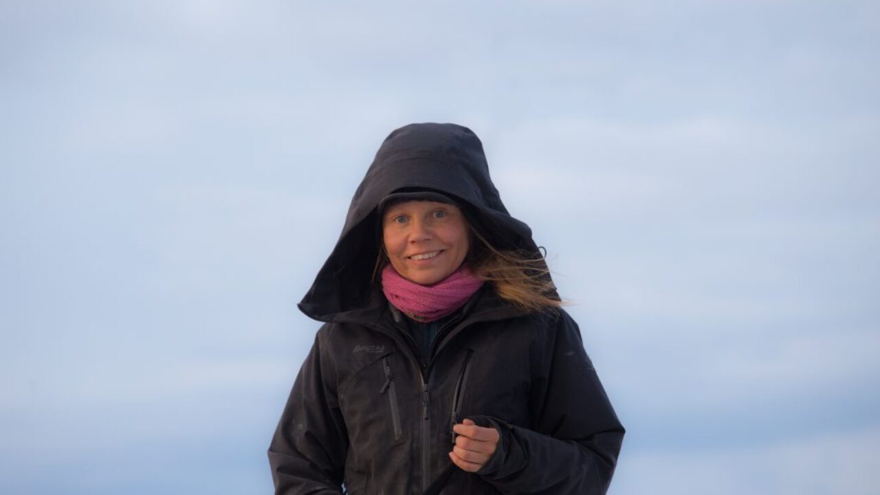 Portrait of Anna Maria standing in the middle of a white landscape. Her light hair is sticking out of the hood of her black jacket and she is smiling broadly. Around her neck is a pink scarf.