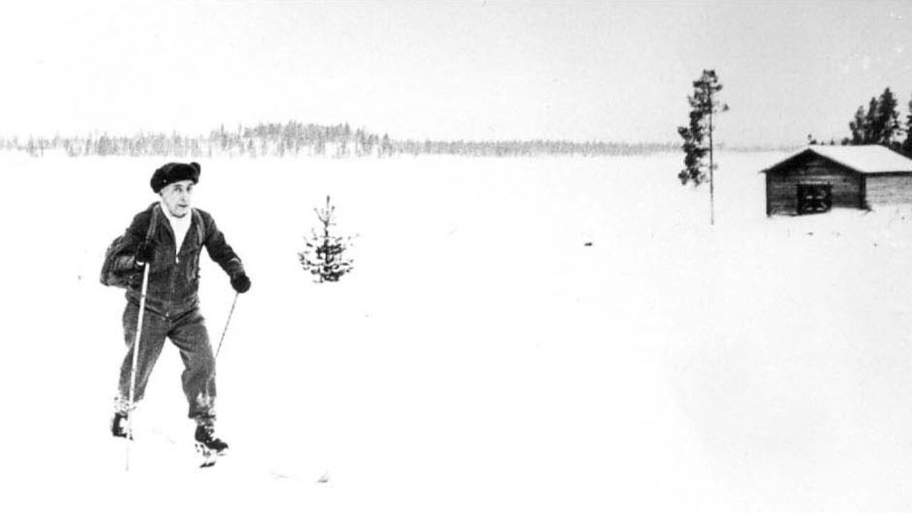 Black and white image of Einar Wallquist skiing in a white winter landscape. Forest in the background and a small timber ring to the right.