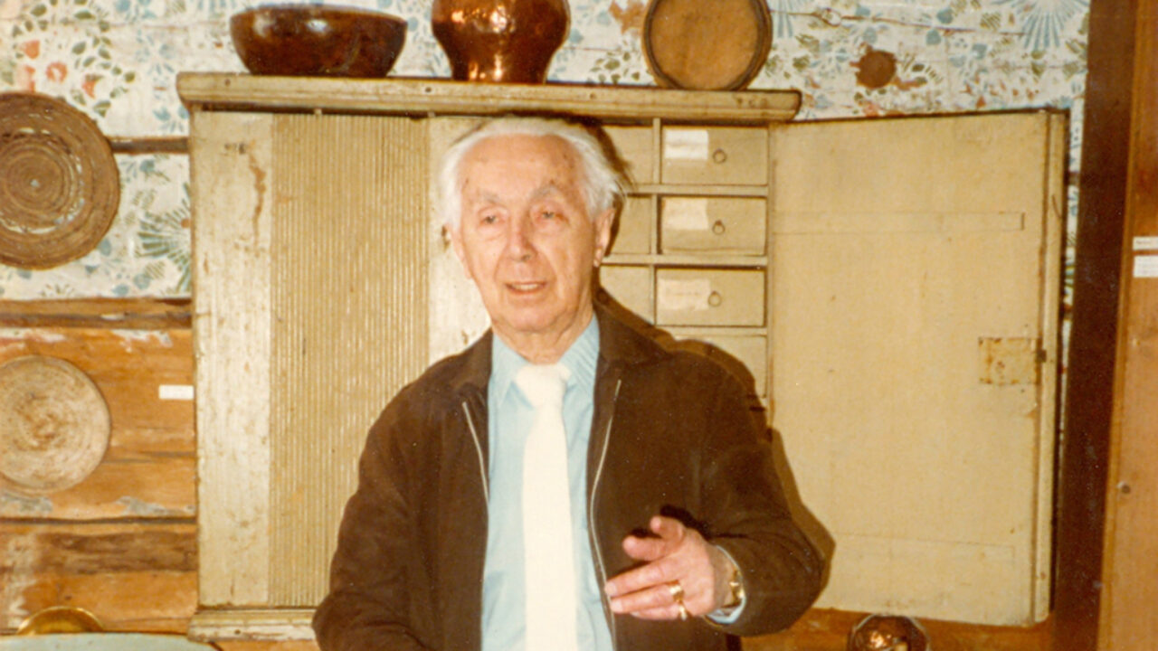 En gråhårig Einar Wallquist i halvfigur framför ett gammalt kryddskåp. Han har grått hår, blå skjorta med vit slips under en brun jacka.