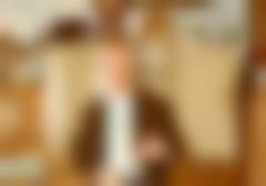 A gray-haired Einar Wallquist in half figure in front of an old spice cabinet. He has gray hair, blue shirt with white tie under a brown jacket.