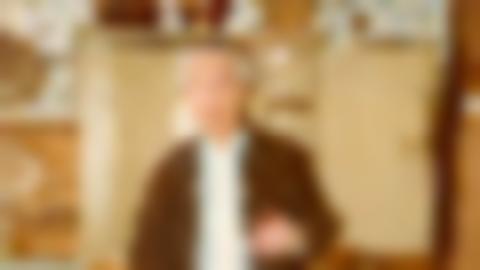 A gray-haired Einar Wallquist in half figure in front of an old spice cabinet. He has gray hair, blue shirt with white tie under a brown jacket.