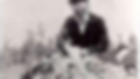 Einar Wallquist sitting on a rock with an open suitcase and painting supplies. He is wearing a beret, glasses and a white shirt under a dark jacket. In the background forest.