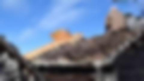 Person med grått, kort hår och gråbrun jacka sitter på taket av en timrad bod. Närmast takets ena nock har det lagts nytt virke, där personen sitter är det näver.