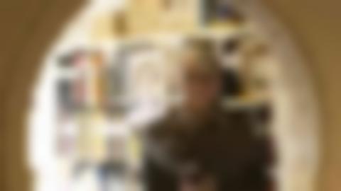 Portrait of Ingela standing holding a binder and looking into the camera through a round window. She has short-cropped gray hair, round glasses and patterned scarf, and a smile on her face. Behind her is a bookshelf.