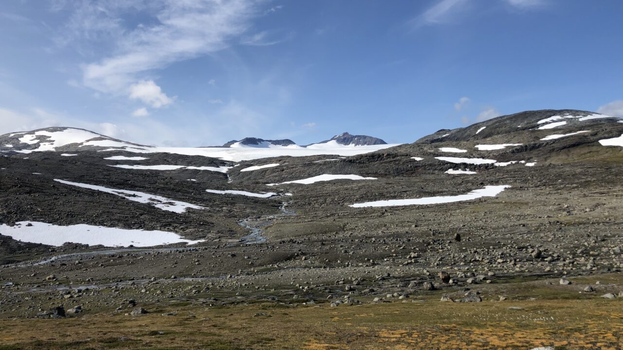 Gråaktig högfjällsmiljö med mycket sten, toppar och snöfläckar. I mitten rinner en bäck.