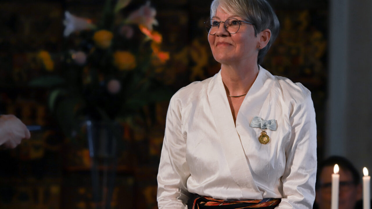 Mot en mörk flerfärgad bakgrund syn Ingela Bergman i vit blus där en medalj i ljusblå rosett är fastsatt. Ingela har kort hår och glasögon.