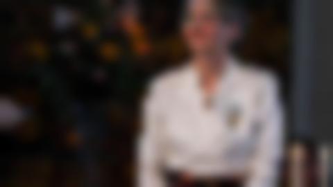 Mot en mörk flerfärgad bakgrund syn Ingela Bergman i vit blus där en medalj i ljusblå rosett är fastsatt. Ingela har kort hår och glasögon.