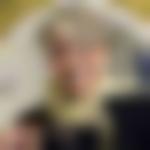 Porträtt av Ingela Bergman. Hon ser leende rakt in i kameran, och håller i sina glasögon. Runt halsen har hon en gul halsduk som hämtar igen färgen den tapet som syns i bakgrunden.