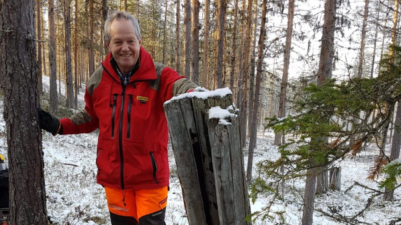 Lars out in the forest, where there is a thin layer of snow on the hill, standing by a gray pine stump. He is wearing a red jacket and orange pants, and he is smiling straight into the camera.