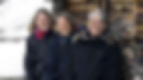 Three smiling women in front of an old log cabin. Gunilla on the far left has semi-long, light hair and glasses and a red scarf, Malin has a dark short page and a turquoise scarf, Ingela on the far right has gray short hair, glasses and a patterned scarf.