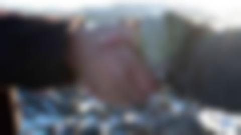 Handshake between two people. One with a brown shirt sleeve and one with a gray shirt sleeve. The sun is shining and there is a rocky landscape with water in the background.