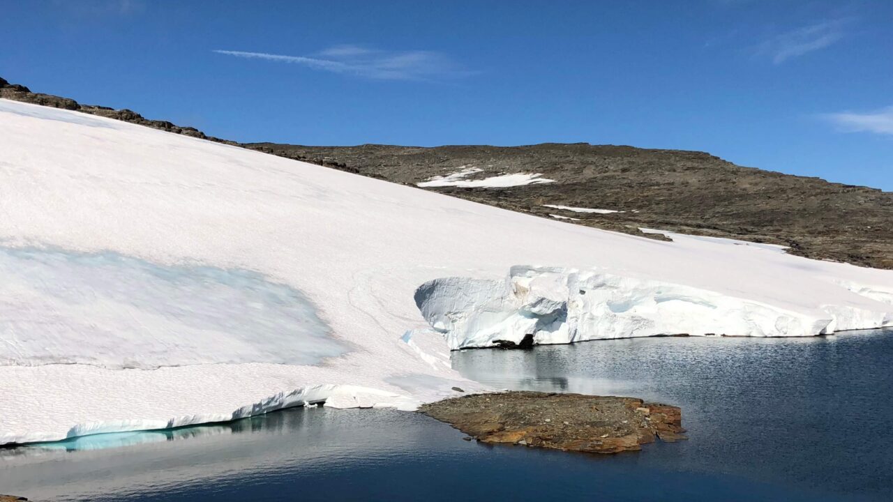 Mitt över bilden sträcker en en bit glaciäris ut sig. Ovanför isen är det klarblå himmel och fjällsluttning, nedanför vatten och grusbacke. I vattnet går det att se en liten del av isen under ytan där den lyser turkos.
