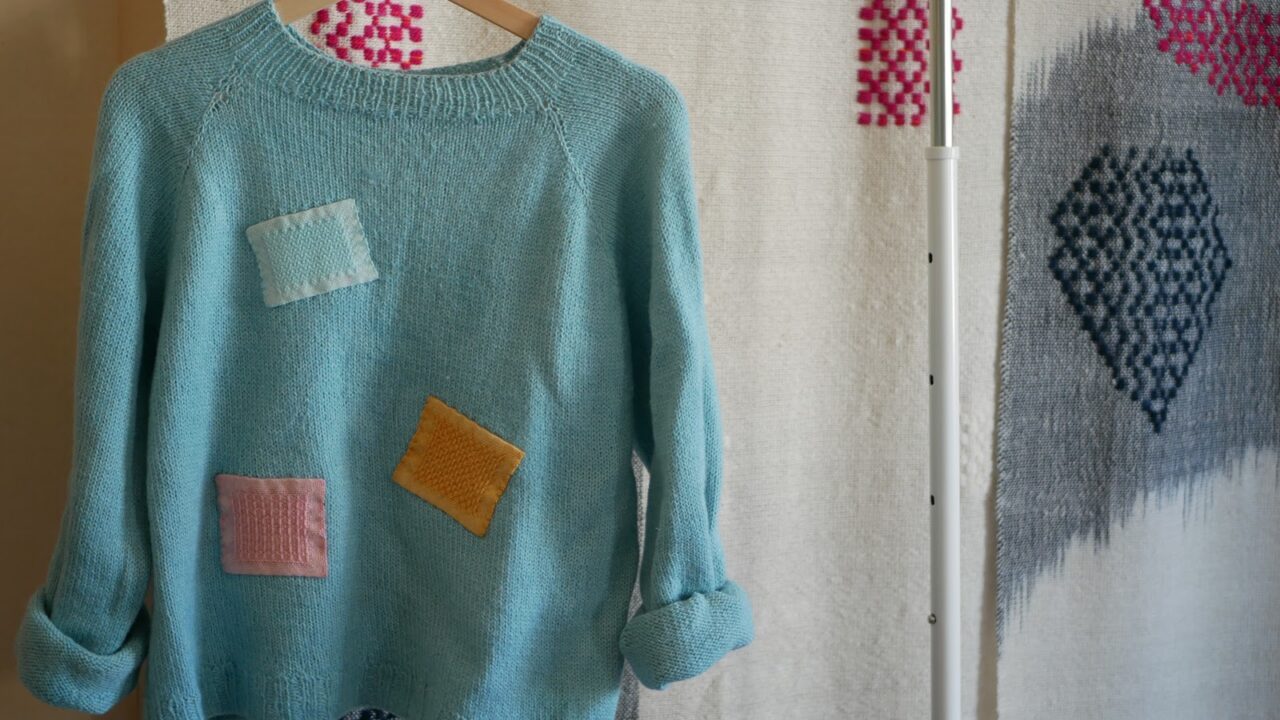 En turkos ylletröja hänger på en galge. Tröjan har tre påsydda "Karra"-lagningslappar i olika färger, rosa, orange och ljusare turkos.