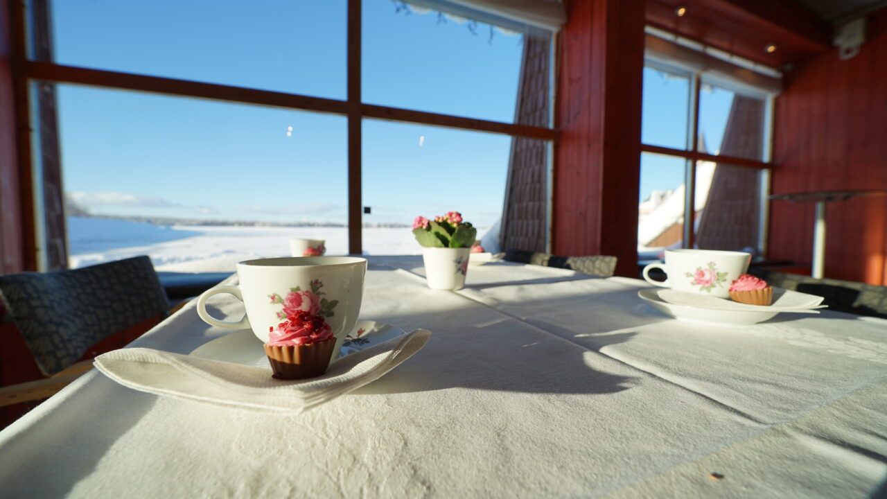 Bord med vit duk och blommiga kaffekoppar framför ett stort fönster. Solen lyser in.