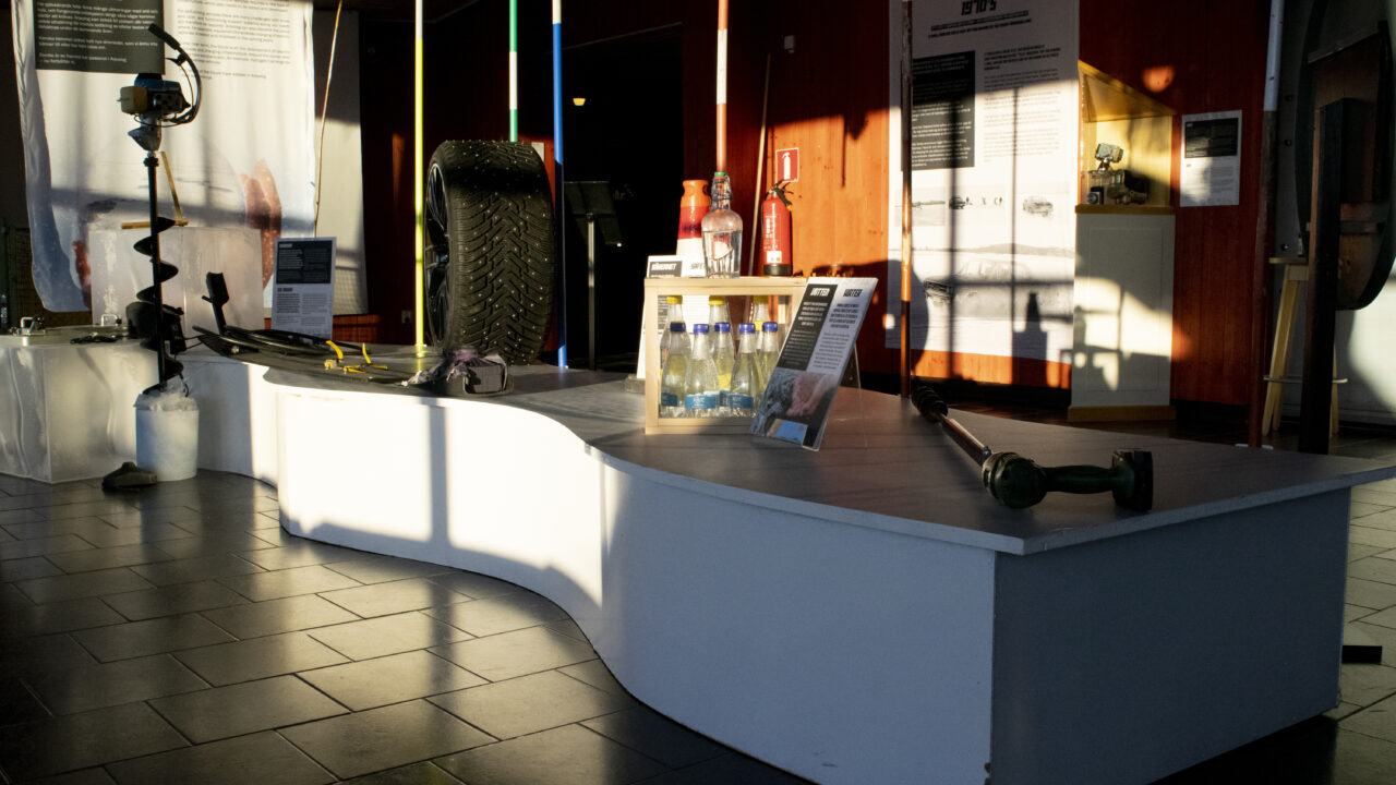 White, curved podium with objects on it, a tire, water bottles, ice drills and road sticks. The sun is shining on the podium, and in the background you can see banners with text and more objects.
