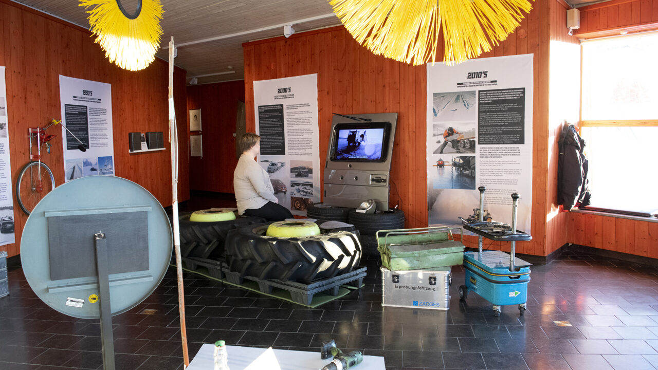 Room with rust-red walls where objects related to the automotive testing industry are displayed. On the walls hang banners with text. On one of two horizontal tractor decks, a person sits and watches a movie shown in the trunk of a car.