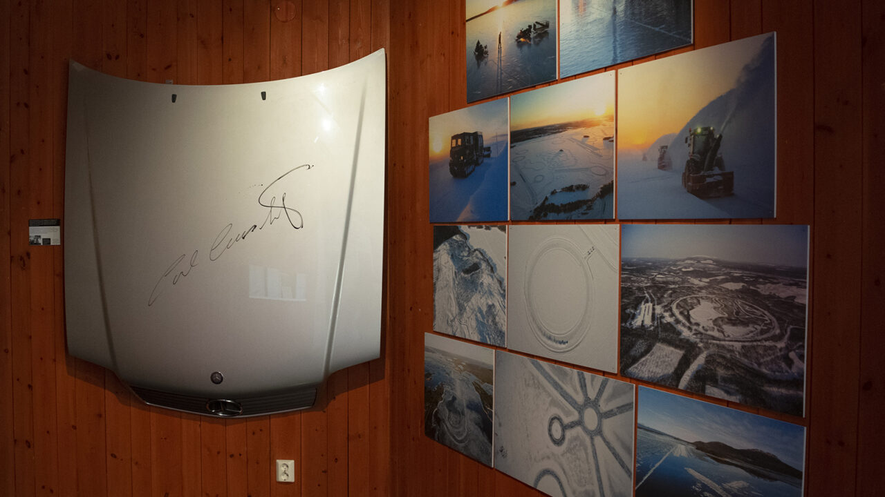 A rust-red wall with a silver bonnet signed by King Carl Gustav. To the right, a wall with photos of test tracks on the ice.