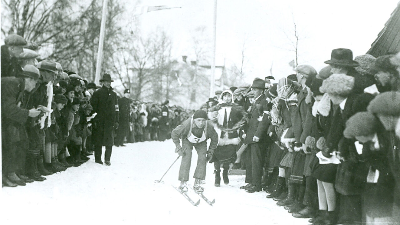 Black and white image of a skier bent forward, skiing forward between a large crowd. Behind the skier a woman is seen running with a wreath.