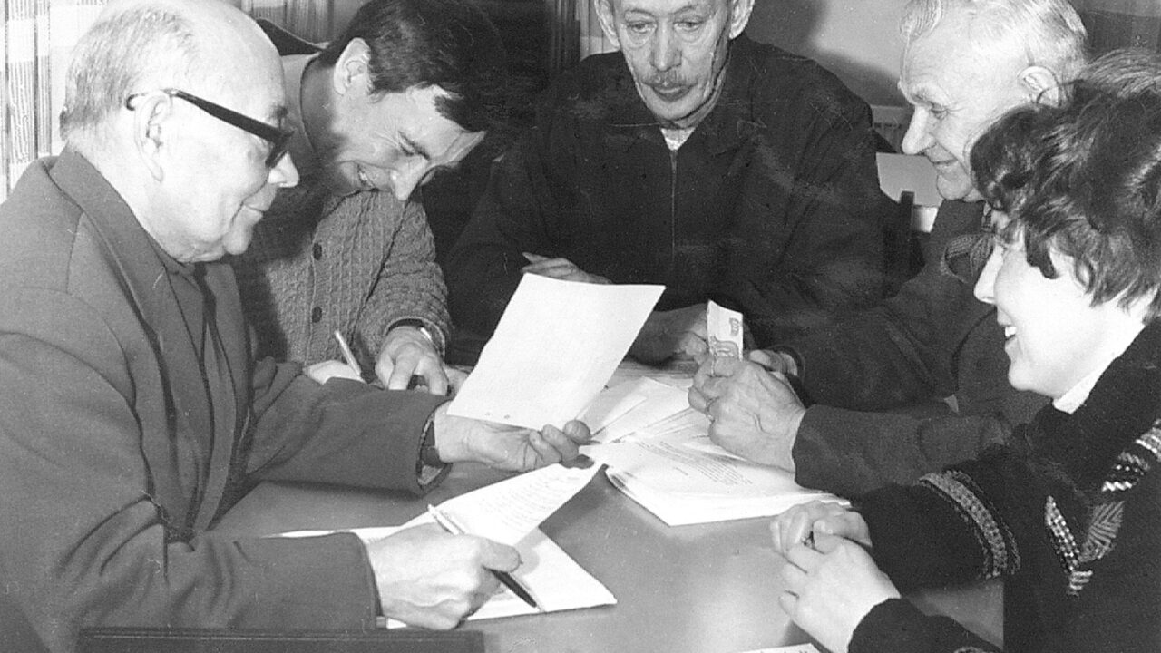 Fyra män och en kvinna sitter runt ett bord med några papper utlagda. De ser glada ut. En skriver på ett papper.