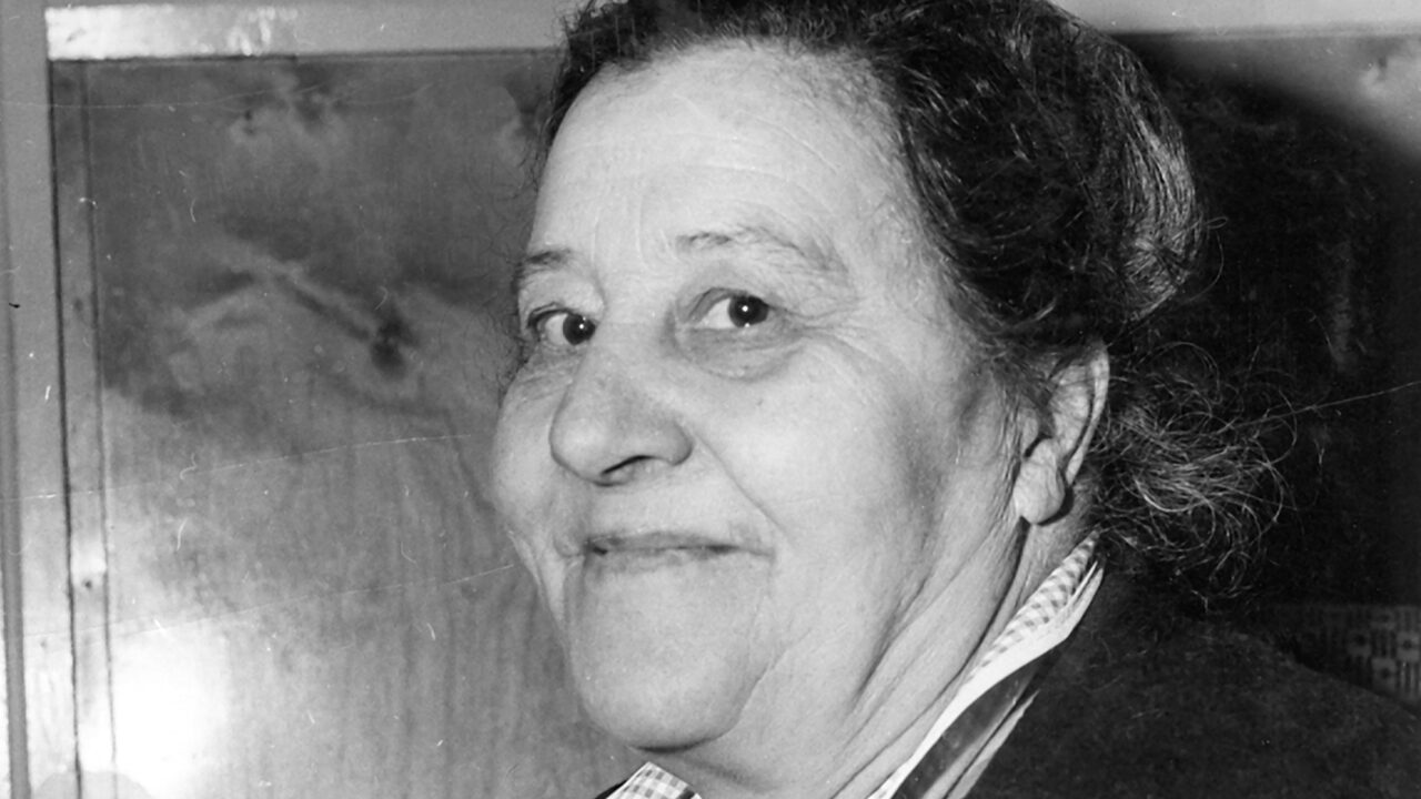 Black and white close-up of older woman with dark hair. The woman looks obliquely into the camera and has a small smile on her lips.