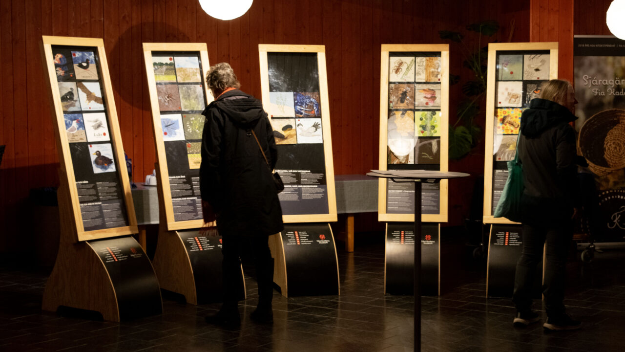 Utställnigslokalen Östan. Roströda väggar och svart stengolv. Fem montrar med broderier står på golvet.