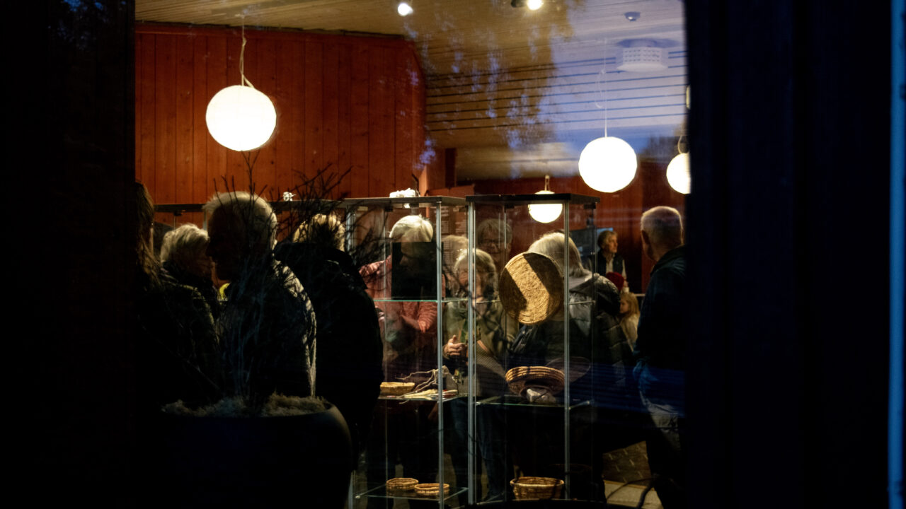 Utställningslokal Östan. Roströda väggar, runda vita taklampor. Bilden tagen utifrån genom fönstret, visar människor som tittar på föremål i montrar.