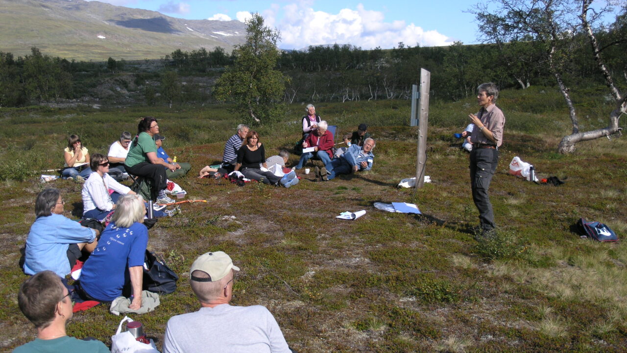 En grupp människor i fjällmiljö sitter på backen och ser mot en person som står upp