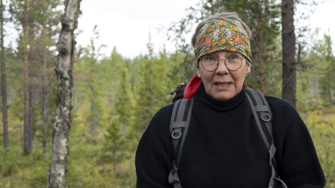 Person med skarf på huvudet, glasögon och svart tröja står i skogen och blickar rakt mot kameran.
