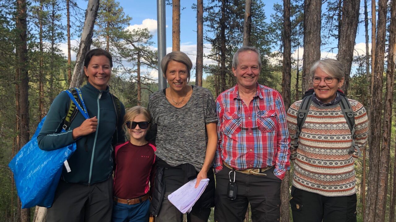 5 personer, 4 vuxna och ett barn, står bredvid varandra och ser leende mot kameran. I bakgrunden skog,