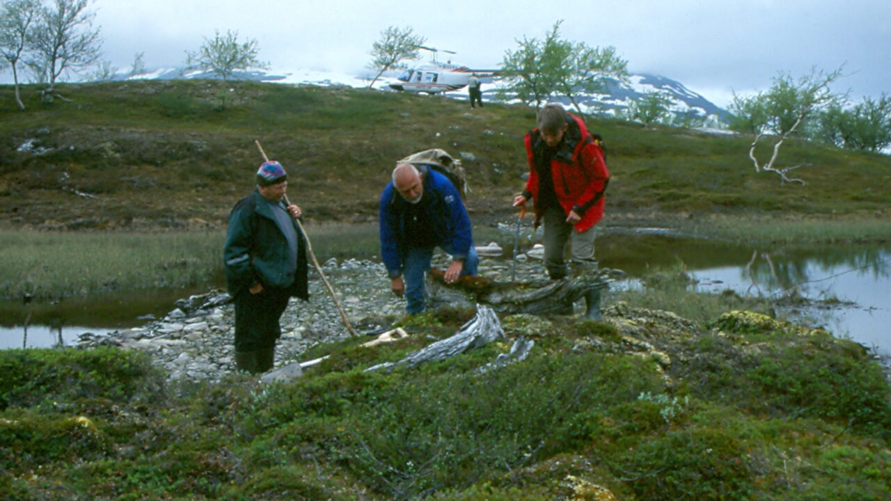 Tre personer i fjällmiljö tittar på något på backen. I bakgrunden syns en helikopter.