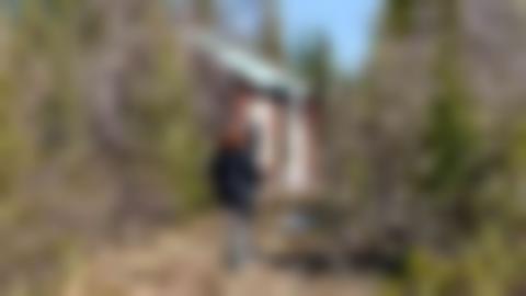 Person med ljusa byxor, svart jacka och rött lockigt hår går på en stig upp mot ett litet timmerhus, Timringen ser sliten och obebott ut.