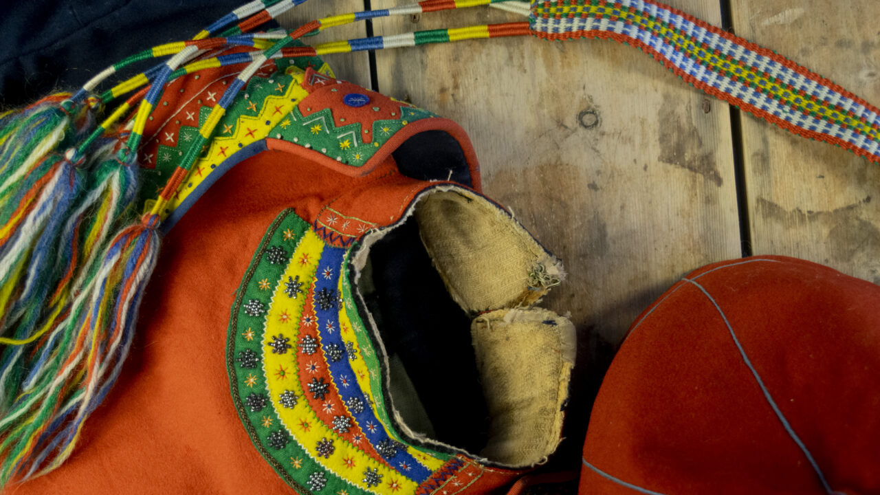 Detaljer på en kvinnokolt som ligger på ett trägolv: barmkläde med röd botten och halvmåneformade delar i grönt, gult rött och blått broderade med pärlor. En röd mössa syns i nedre högra hörnet, en del av ett vävt bälte i bildens ovandel