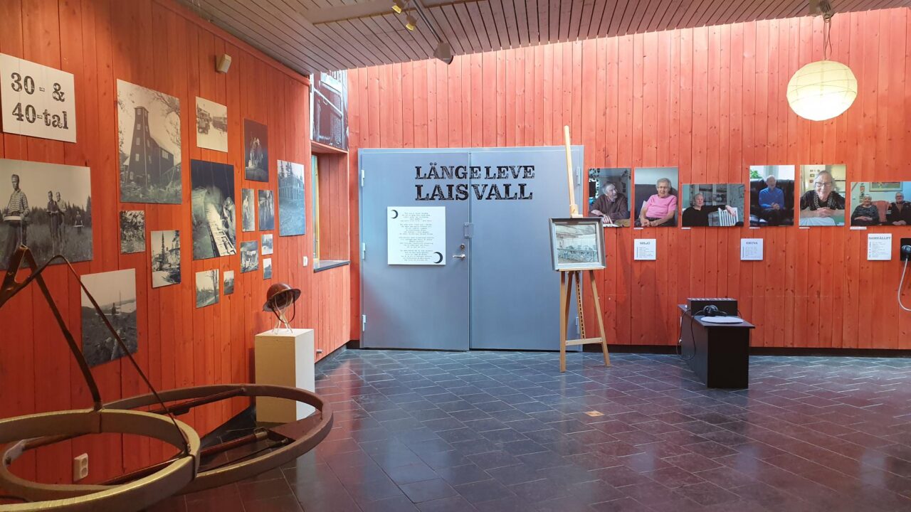 Parti ur utställningen. Fotografier på roströd vägg. Stor text "Länge leve Laisvall" på en grå dörr